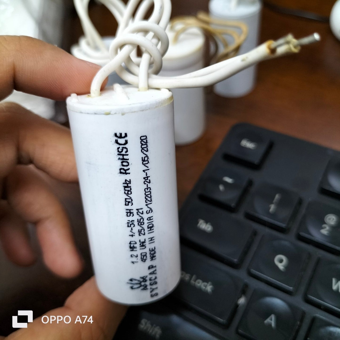 Hand holding a white Motor Running Capacitor 1.2MFD 450v with wires near a black keyboard