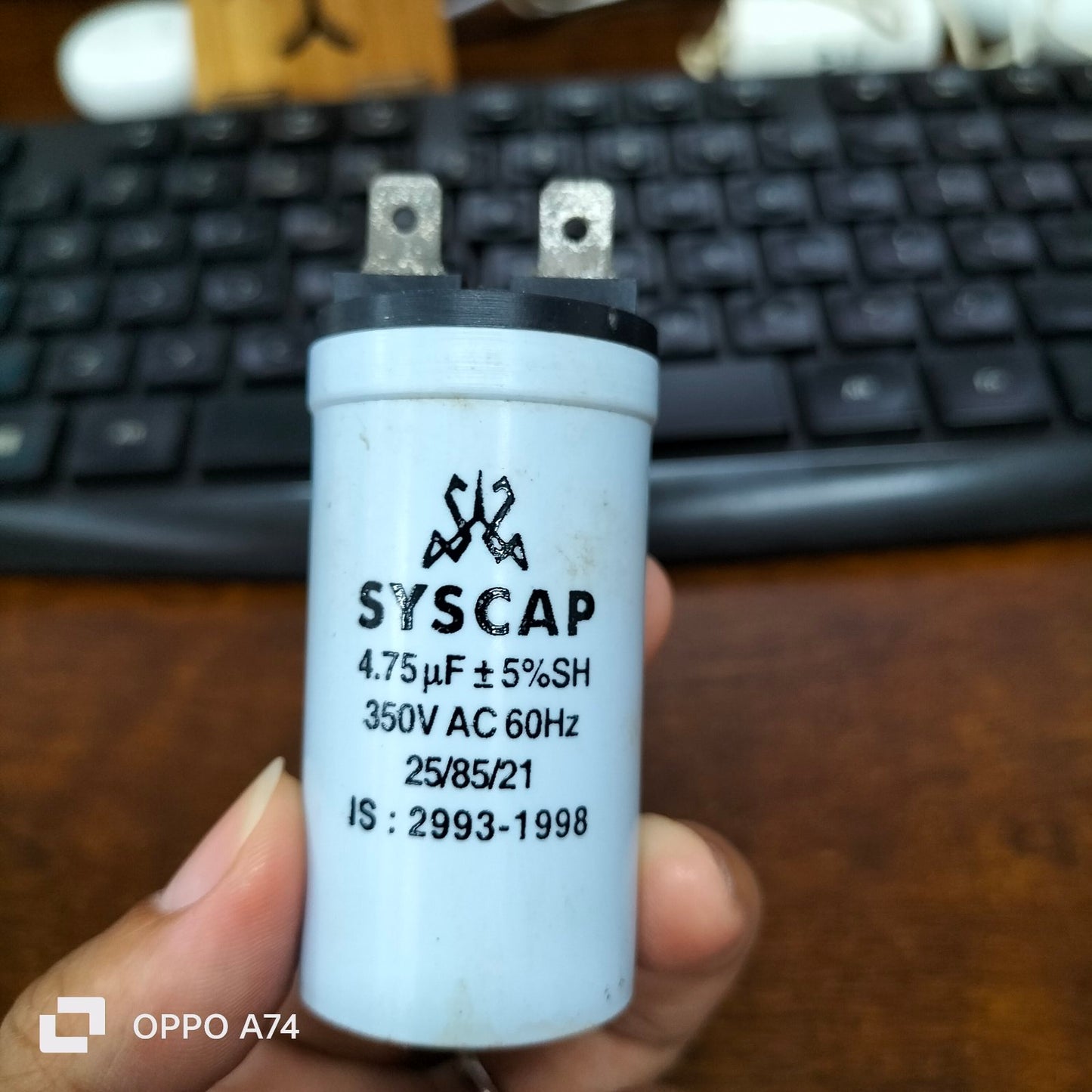 Syscap Running Capacitor 4.75MFD 350 VAC electrical component held in hand with black keyboard in background
