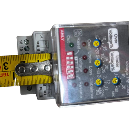 A close-up of a voltage trip dial indicator or electrical monitoring module with a clear plastic housing, silver internal components, red and yellow labels, and a yellow tape measure being held against it on a white background.