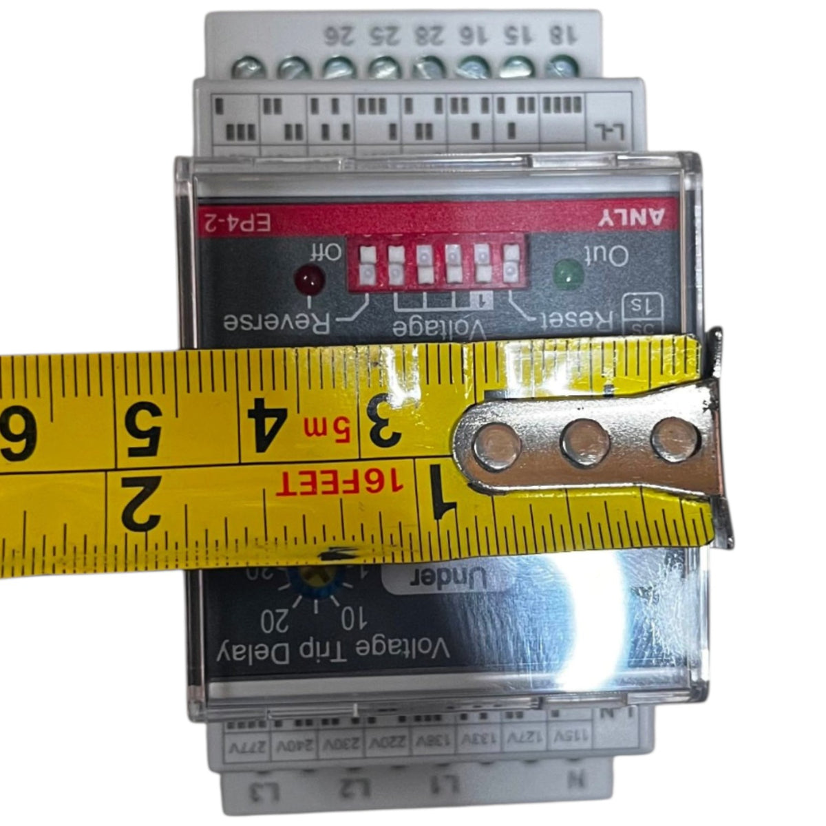A voltage trip delay electrical relay module with a clear plastic housing, showing red and green indicator lights, terminal blocks on top and bottom, and a yellow tape measure laid horizontally across the front to indicate size.