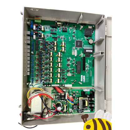 An open metal electrical control cabinet showing a large green circuit board packed with electronic components, capacitors, and wiring, photographed on a white background with a small bee graphic in the bottom right corner.