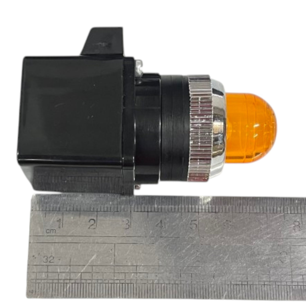 A small black electrical switch module with a clear amber/orange dome light and a silver ribbed metal collar, shown next to a metal ruler for scale on a white background.
