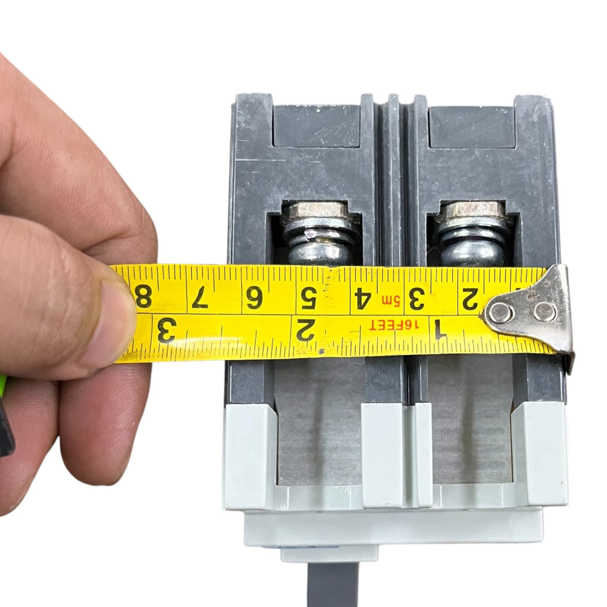 A close-up of a hand holding a yellow tape measure against a gray electrical terminal block to show its width in centimeters and inches.