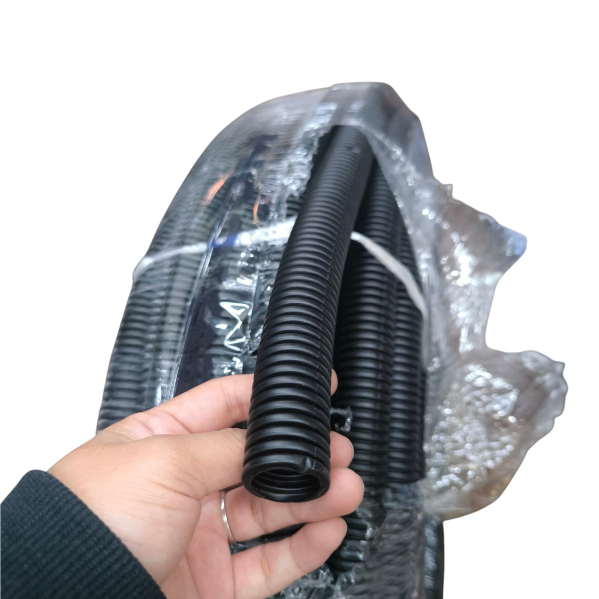 A hand holding a short section of black corrugated plastic conduit tube, with a larger spool of the same tube wrapped in clear protective plastic in the background.