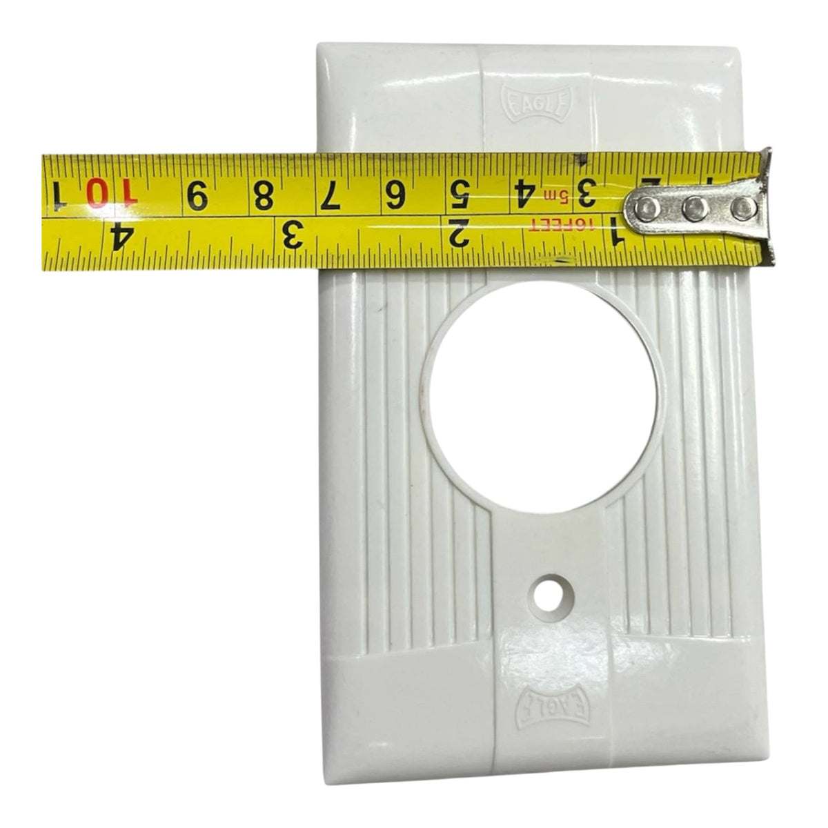 A white plastic outlet cover plate with a large circular cutout and a small round screw hole, shown with a yellow tape measure stretched horizontally across the top edge to indicate size.