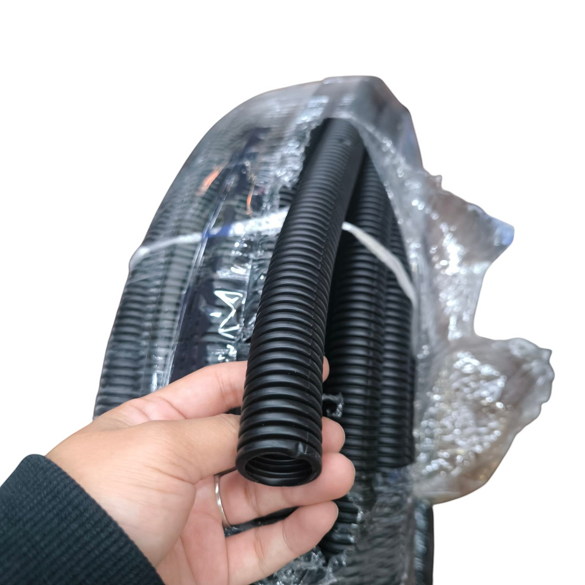 A hand holding a black corrugated plastic conduit tube, with a larger coil of the same black tubing wrapped in clear protective plastic in the background.
