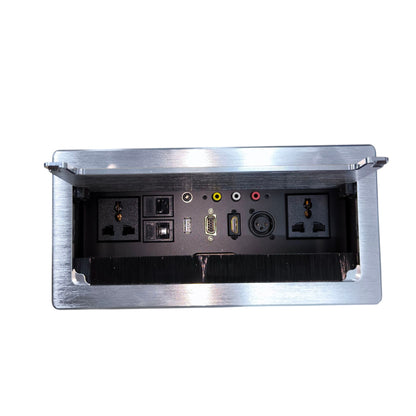 A brushed stainless steel under-desk power outlet cover with its lid open, revealing multiple electrical outlets, switches, and audio/video connection ports on a black interior panel, isolated on a white background.