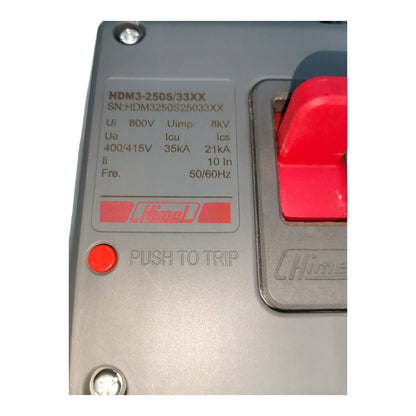 Close-up of a Hima electrical component, showing the model HDM3-250S/33XX, technical specifications on a silver metal faceplate, a red push-to-trip button, and part of a red switch.