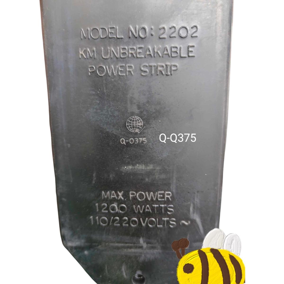 A close-up of a dark gray metal power strip component with embossed text reading 'MODEL NO: 2202 KM UNBREAKABLE POWER STRIP' and 'MAX POWER 1200 WATTS 110/220VOLTS ~'. A small bee graphic appears in the bottom right corner.