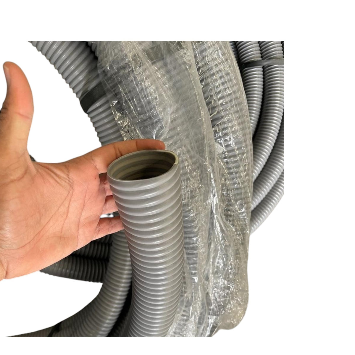 A hand holding a gray corrugated plastic pipe fitting in front of a coiled bundle of similar gray flexible pipes wrapped in clear protective plastic, on a white background.