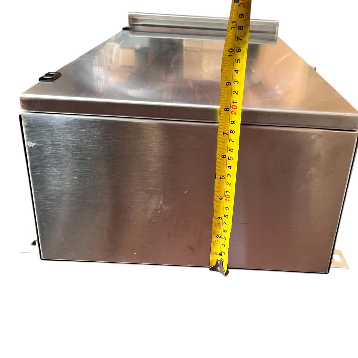 A stainless steel rectangular metal box with a lid, shown against a white background with a yellow tape measure standing vertically to indicate its height.