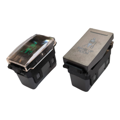 Two automotive toggle switches on a white background, one with a clear cover showing a green illuminated symbol and the other with a gray textured top and 'MANSET UP BEFORE' text.