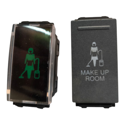 Two industrial toggle switches side by side on a white background. The left switch has a clear cover showing a green illuminated symbol of a person with a vacuum cleaner.