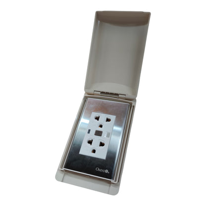 An outdoor waterproof electrical outlet cover with an open hinged lid, showing a white three-pin socket protected by a clear cover over a brushed stainless steel faceplate. The brand name 'Outdoor' is printed at the bottom of the plate.