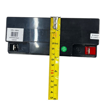 A 12V 18Ah black battery shown from the back with a yellow tape measure vertically aligned to display its height. The battery has white labels on both sides, a green circular sticker, and a red terminal cover on the right.