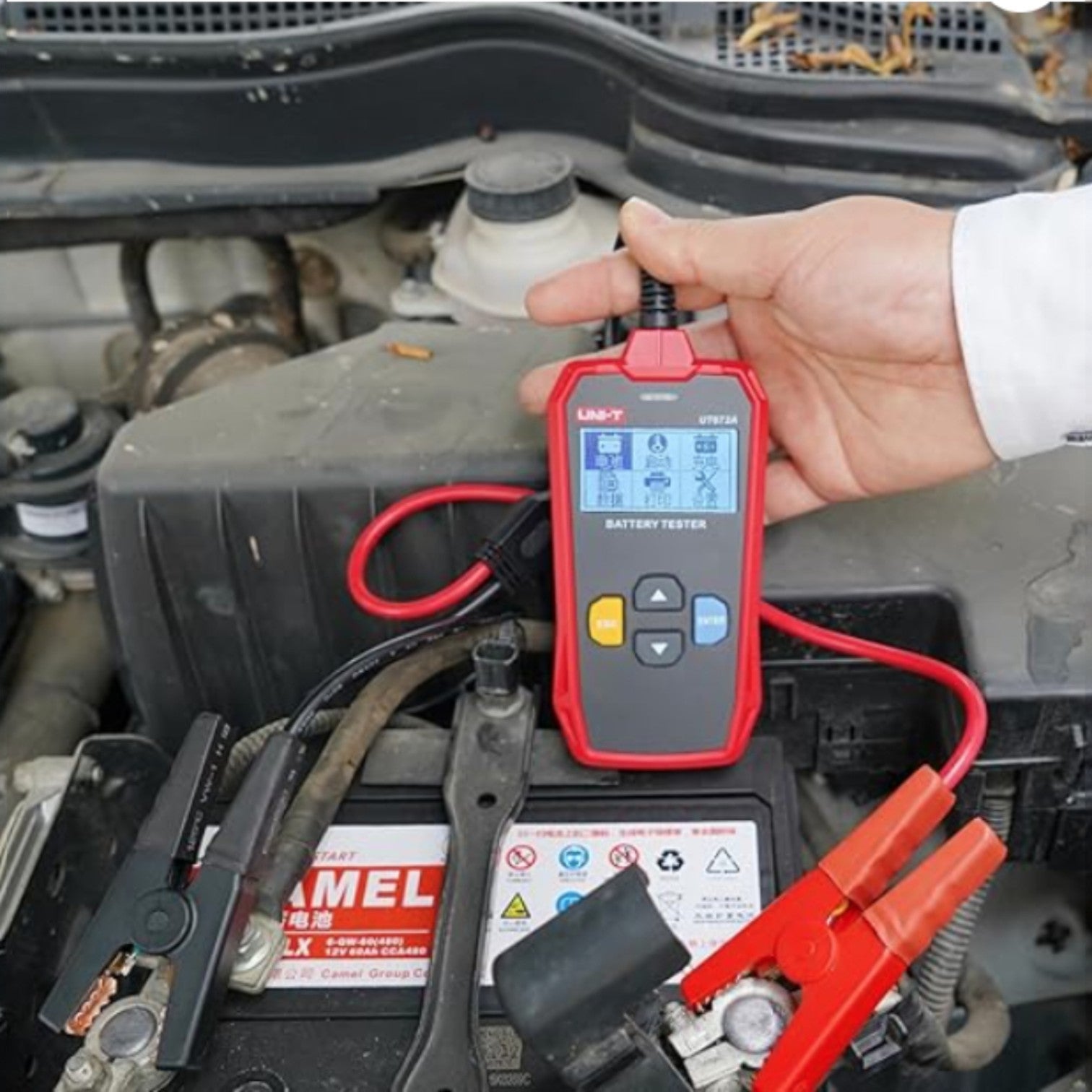 Person using a red battery tester on a car battery with a clear view of the engine compartment. UNI-T Digital Battery Tester 12V or 24V 250AH Max Capacity UT673A Electx Electrical Supply